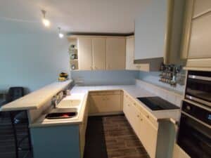 Cuisine moderne lumineuse avec des meubles beiges, des comptoirs carrelés blancs, un double évier, une plaque de cuisson et des fours intégrés. Un tabouret de bar est visible sur un sol carrelé foncé.