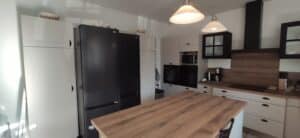 Cuisine moderne avec meubles blancs, grand réfrigérateur LG noir, îlot central et plan de travail en bois. Électroménager intégré, hottes et luminaires suspendus.