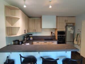 Cuisine moderne avec armoires en bois clair, plans de travail et îlot bar gris marbré. Équipée d'un four, micro-ondes, réfrigérateur et plaques induction. Trois tabourets de bar noirs sont visibles.