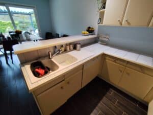 Cuisine moderne beige et bleue avec double évier blanc, carrelage clair, lave-vaisselle Bosch intégré. Grande fenêtre donnant sur un jardin verdoyant.