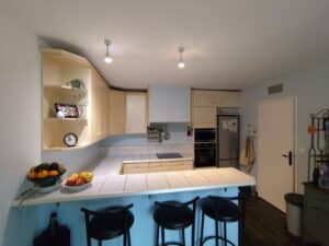 Cuisine moderne avec îlot central carrelé, tabourets noirs, meubles crème et plans de travail blancs. Four encastré, frigo, fruits frais, horloge.