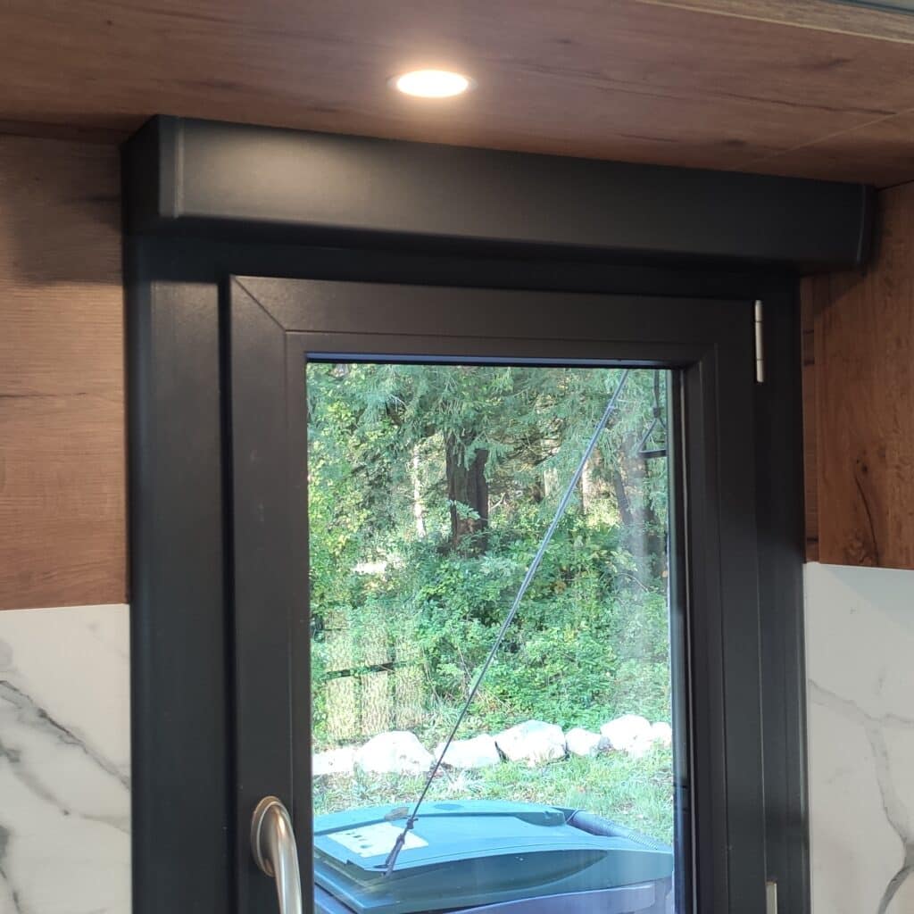 Fenêtre/porte moderne noire avec store intégré. Vue sur forêt luxuriante. Plafond en bois avec spot allumé et comptoir marbre blanc.