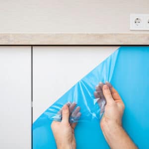 Mains retirant un film protecteur bleu d'un meuble de cuisine blanc. La surface lisse apparaît sous le plan de travail et des prises.