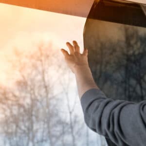 Main posant un film de protection solaire sur une fenêtre, la partie non couverte laisse passer une lumière chaude orangée et révèle des arbres flous.